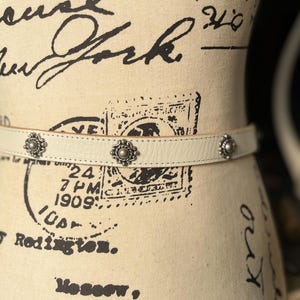 May include: Cream leather belt with three silver floral studs. The belt is displayed on a mannequin with vintage text and stamps. A fashionable accessory for trousers or skirts.