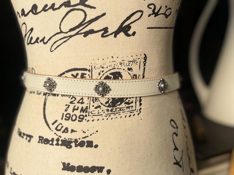 May include: Cream leather belt with three silver floral studs. The belt is displayed on a mannequin with vintage text and stamps. A fashionable accessory for trousers or skirts.