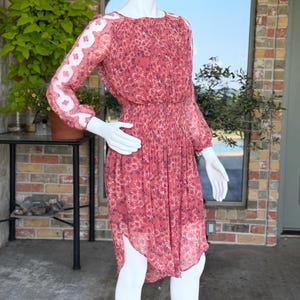 Pode incluir: Um vestido vermelho com estampa floral, mangas compridas e saia plissada. O vestido apresenta um decote redondo e uma cintura franzida. As mangas t&ecirc;m um detalhe em renda branca. O vestido &eacute; exibido em um manequim.