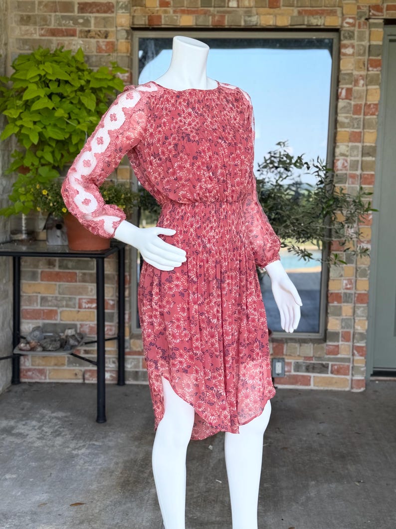 Pode incluir: Um vestido vermelho com estampa floral, mangas compridas e saia plissada. O vestido apresenta um decote redondo e uma cintura franzida. As mangas t&ecirc;m um detalhe em renda branca. O vestido &eacute; exibido em um manequim.