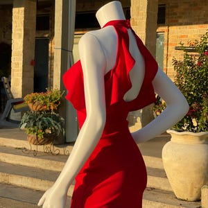 May include: A vibrant red dress with a halter neckline and a ruffled back detail. The dress is displayed on a mannequin, showcasing its form-fitting silhouette. The dress is a solid color, perfect for a special occasion.