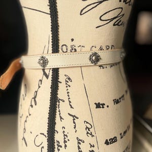 May include: Cream leather belt with silver floral studs. The belt is fastened with a small, tan leather loop. The belt is displayed on a mannequin with script writing.
