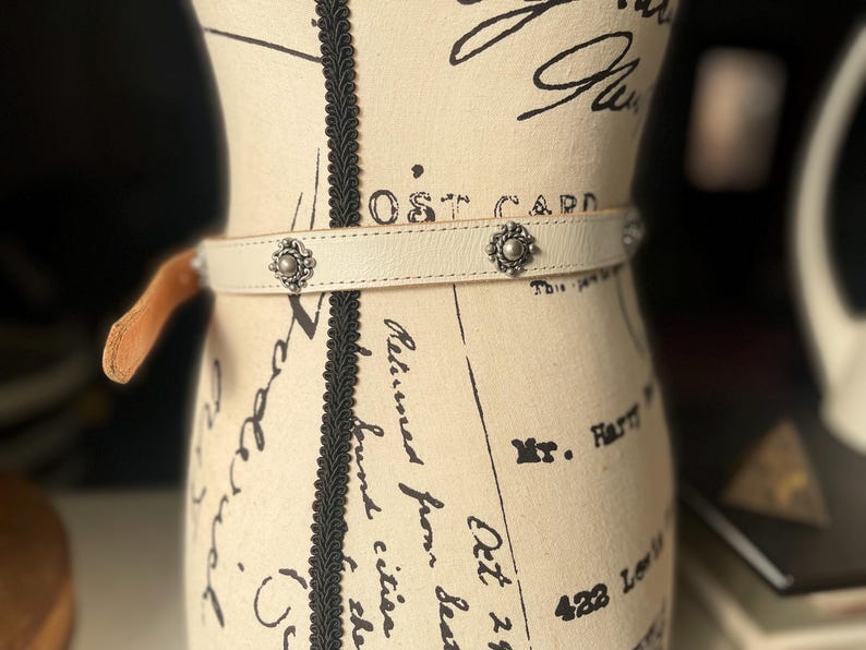 May include: Cream leather belt with silver floral studs. The belt is fastened with a small, tan leather loop. The belt is displayed on a mannequin with script writing.