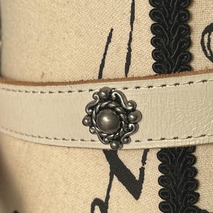 May include: Cream leather belt with silver metal studs. The belt has a decorative central piece and smaller studs. The stitching is visible along the edges. A stylish accessory, suitable for various outfits.