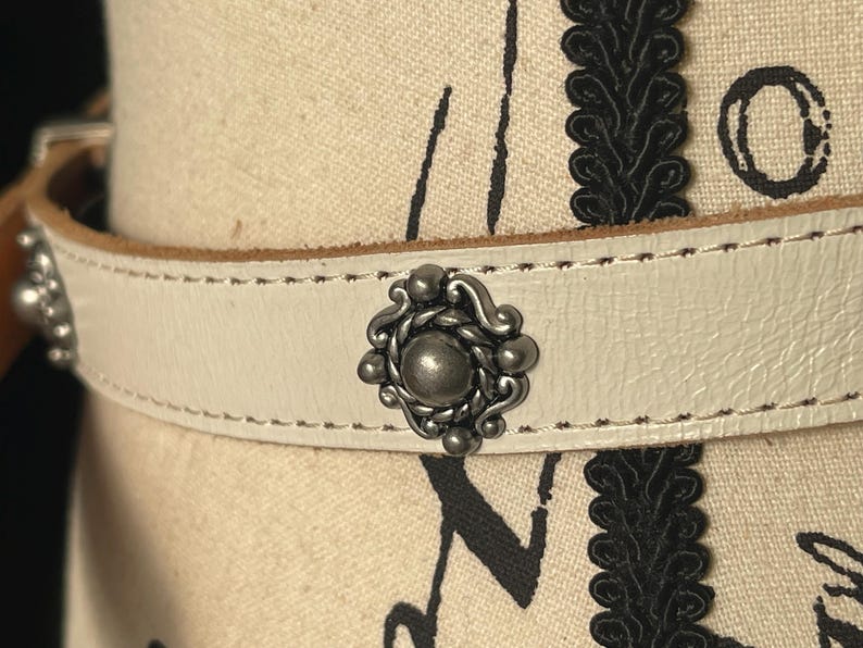 May include: Cream leather belt with silver metal studs. The belt has a decorative central piece and smaller studs. The stitching is visible along the edges. A stylish accessory, suitable for various outfits.