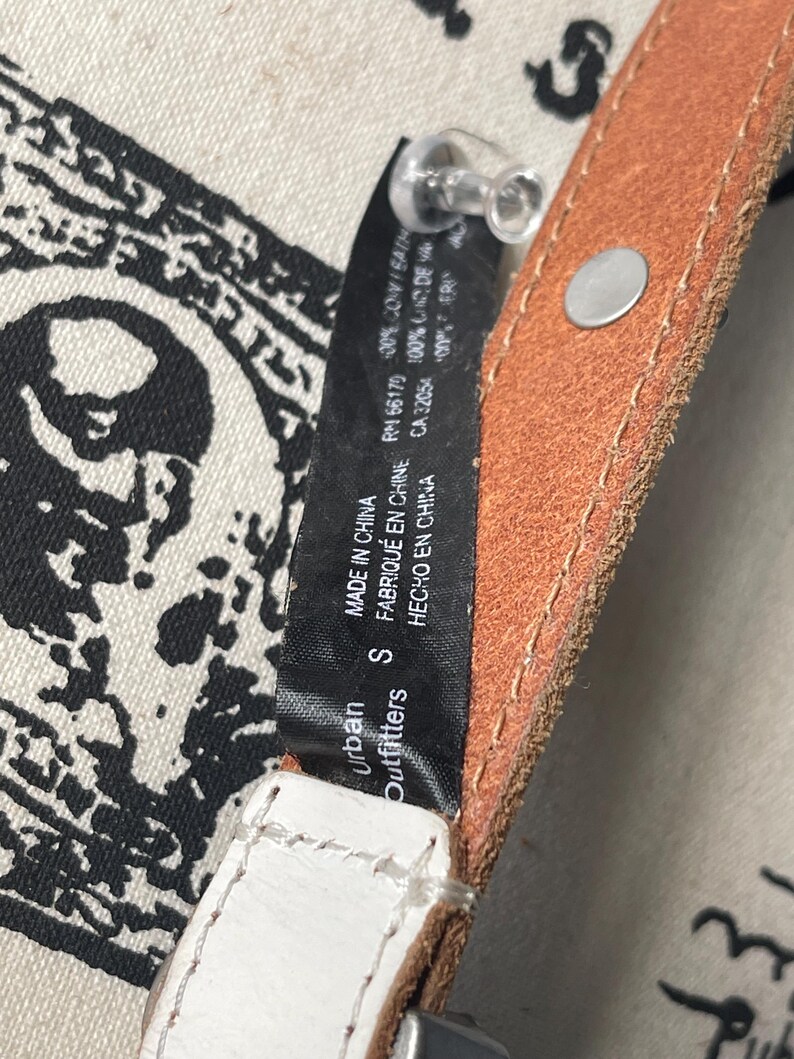 May include: Close-up of a brown and white leather belt with a silver buckle. The belt has a small tag that reads "Urban Outfitters" and "MADE IN CHINA". The belt is a size small.