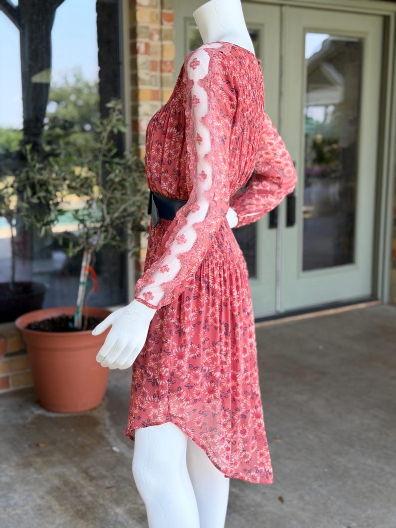 Pode incluir: Vestido de manga comprida com estampa floral em tons de vermelho e rosa, preso na cintura com um cinto preto. O vestido apresenta mangas transparentes com detalhes em renda branca. O manequim est&aacute; usando leggings brancas.