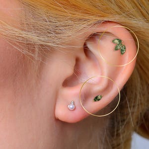 May include: Close-up of an ear with multiple earrings. A small pearl stud is in the lobe, a tiny green stud is in the mid-ear, and a cluster of green leaf-shaped earrings are in the upper ear.