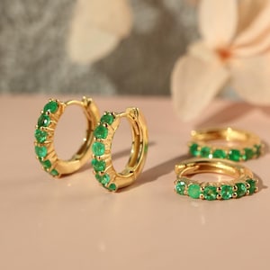 May include: A pair of gold hoop earrings featuring a row of small, round, vibrant green gemstones set along the front curve. The earrings have a polished metallic finish and a hinged closure mechanism, showcasing a bright and reflective surface.