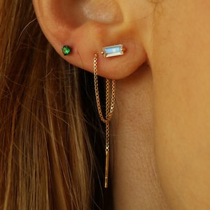 Peut inclure: Gros plan sur une oreille ornée de trois boucles d'oreilles. Un petit clou en diamant, un clou en pierre verte et une boucle d'oreille en chaîne dorée avec une pierre rectangulaire irisée. La boucle d'oreille en chaîne pend avec une barre verticale.