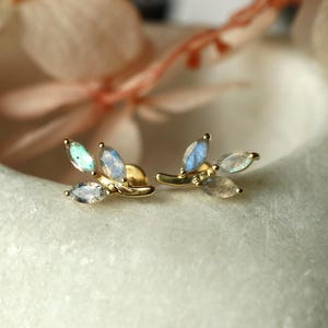 May include: A pair of gold stud earrings. Each earring has a leaf design with three marquise-cut gemstones in blue, green, and clear. The earrings are set against a soft, blurred background.