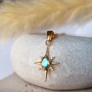 May include: A gold pendant necklace with a starburst design. The pendant features an oval, blue-green gemstone at its centre, encircled by small, clear stones. The starburst has pointed rays extending outwards.