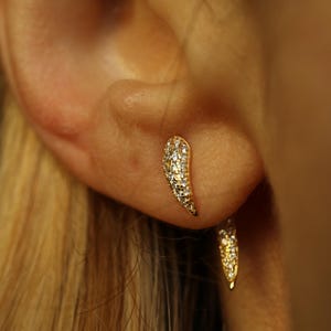 Pode incluir: Brincos dourados com design curvo em forma de chifre, incrustados com pequenos cristais brilhantes. A imagem em close-up destaca os detalhes e como os brincos capturam a luz, criando um efeito deslumbrante.