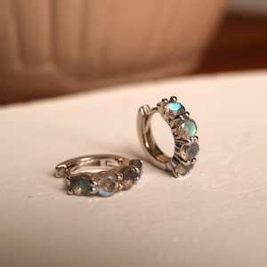 Peut inclure: Deux boucles d'oreilles créoles argentées, chacune ornée de cinq pierres précieuses rondes et irisées. Les pierres précieuses présentent une gamme de couleurs, dont le bleu, le vert et le gris. Les boucles d'oreilles sont présentées sur une surface blanche.