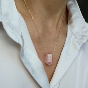 Può includere: Una delicata collana a catena dorata con un ciondolo geometrico sfaccettato rosa pallido.