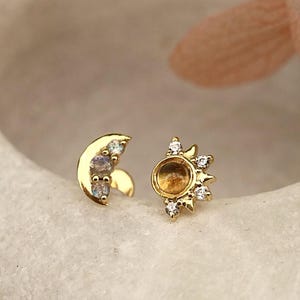 Op de afbeelding: Een paar oorbellen met een hemels thema. Eén oorbel is een gouden halve maan met kleine, iriserende stenen. De andere is een gouden zonnepatroon met een centrale amberkleurige steen en omringende heldere kristallen.