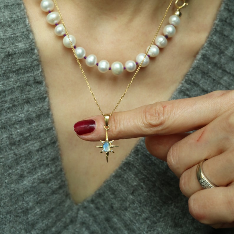 Puede incluir: Un collar de oro con un colgante en forma de estrella con una piedra preciosa azul y peque&ntilde;os diamantes. Tambi&eacute;n se ve un segundo collar de perlas blancas con hilo morado. El fondo es un su&eacute;ter gris.