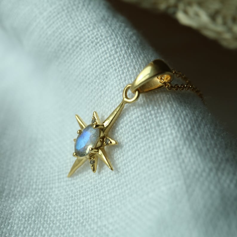 Puede incluir: Un collar con colgante de estrella dorada con una piedra preciosa ovalada e iridiscente en el centro. La estrella tiene ocho puntas y peque&ntilde;as piedras de acento. La delicada cadena dorada est&aacute; sobre un fondo de tela blanca.