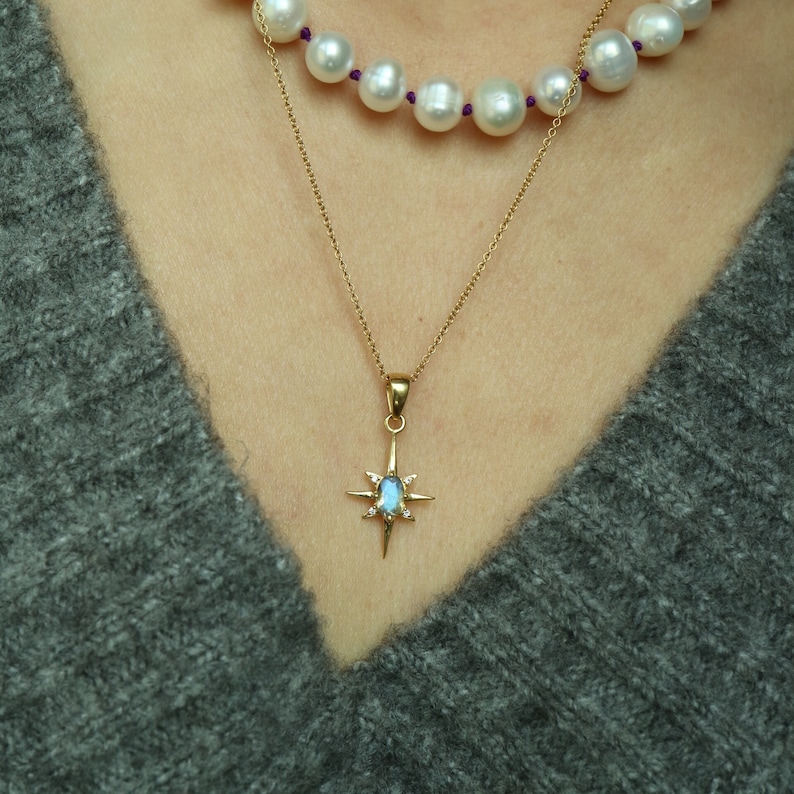Puede incluir: Un collar de oro con un colgante en forma de estrella con una piedra preciosa azul y peque&ntilde;as piedras blancas. Tambi&eacute;n es visible un segundo collar de grandes perlas blancas con detalles morados. Los collares se llevan sobre un jersey gris.