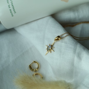 Puede incluir: Un collar dorado con un colgante en forma de estrella y un par de peque&ntilde;os pendientes de aro dorados. El colgante tiene una piedra central. Un tubo de tratamiento de manos est&aacute; en el fondo. Las joyas est&aacute;n sobre tela blanca.