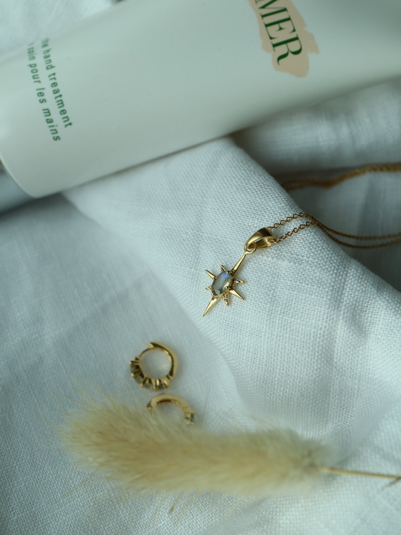 Puede incluir: Un collar dorado con un colgante en forma de estrella y un par de peque&ntilde;os pendientes de aro dorados. El colgante tiene una piedra central. Un tubo de tratamiento de manos est&aacute; en el fondo. Las joyas est&aacute;n sobre tela blanca.
