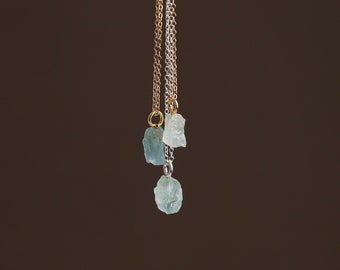 May include: Three delicate necklaces with raw aquamarine stone pendants hanging from gold and silver chains.