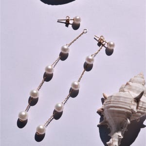 Puede incluir: Un par de pendientes de oro con perlas blancas. Los pendientes están sobre un fondo blanco con conchas.