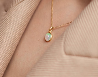 May include: A delicate gold chain necklace with a small oval opal pendant. The opal is iridescent with shades of white, green, and blue.