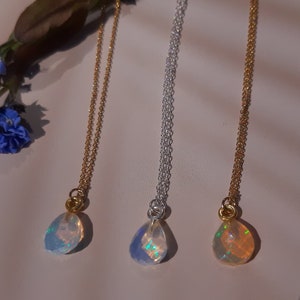Op de afbeelding: Drie delicate kettingen met traanvormige opaal hangers. De kettingen liggen op een wit oppervlak. De hangers zijn iriserend en hebben een regenboog aan kleuren.