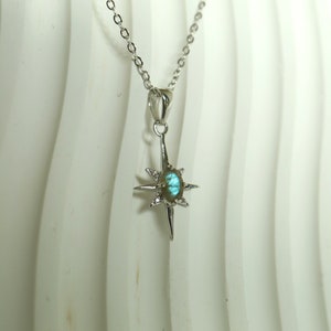 Puede incluir: Un collar de plata con un colgante en forma de estrella. El colgante tiene una piedra preciosa ovalada iridiscente en el centro, rayos puntiagudos y peque&ntilde;as piedras claras. La delicada cadena es de plata.