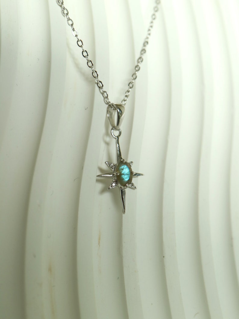 Puede incluir: Un collar de plata con un colgante en forma de estrella. El colgante tiene una piedra preciosa ovalada iridiscente en el centro, rayos puntiagudos y peque&ntilde;as piedras claras. La delicada cadena es de plata.