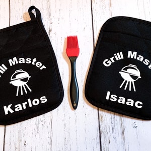May include: Two black oven mitts with white text that reads "Grill Master" and the names "Karlos" and "Isaac". A red silicone basting brush is in the center of the image.