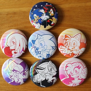 May include: Seven circular pin badges featuring characters from the Sonic the Hedgehog series. The badges showcase Sonic, Shadow, Knuckles, and Tails in different designs and colours. The badges are arranged on a wooden surface.