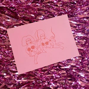 May include: A pink greeting card featuring a line drawing of two cartoon women in a relaxed pose. The women are wearing glasses, and their bodies are partially covered. The card is set against a background of pink tinsel.