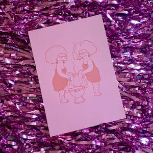 May include: A pink art print featuring two cartoon women with large afros, wearing dresses, and smoking cigarettes while sitting on a toilet. The illustration is in a peach color. The background is a shiny, pink, tinsel-like material.