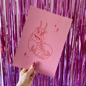 May include: A pink print with a cartoon lobster with a smiling face and a pink background. The lobster is outlined in red and has a few small stars around it.