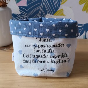 Peut inclure: Un petit panier de rangement en tissu avec une bordure bleue à motifs de cœurs et une base blanche. Le panier porte une citation en français, attribuée à Walt Disney. Le fond présente un motif floral.