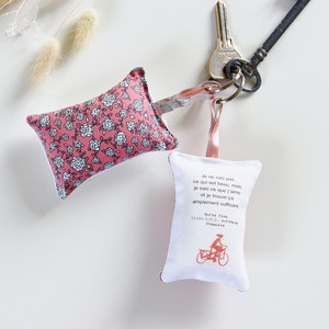 May include: A pink floral fabric keychain with a small white fabric pouch attached. The pouch has a quote in French and a bicycle illustration. The keychain is attached to a silver key ring and a vintage-style key.