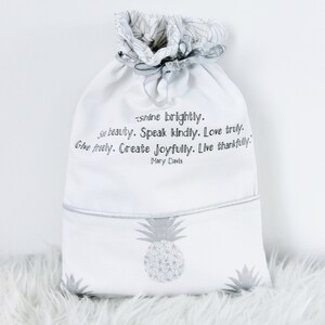 May include: White drawstring bag with a pineapple print and the quote "Shine brightly. See beauty. Speak kindly. Love truly. Give freely. Create joyfully. Live thankfully." by Mary Davis.
