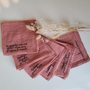 Op de afbeelding: Set van zes roze muslin servetten met zwarte geborduurde Franse zinnen. De zinnen zijn: "Se raconter nos vies autour d'un verre et refaire le monde", "Partager, déguster, se régaler", "Partager un bon repas", "Goûter aux plats nouveaux", "Chanter et rire jusqu'à pas d'heure" en "Cuisiner avec amour".