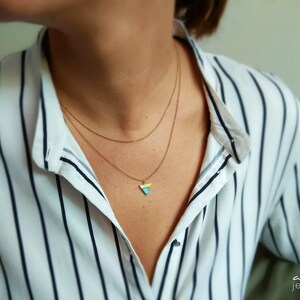 May include: A gold necklace with a small triangle pendant. The pendant is made of yellow, blue, and white enamel. The necklace is layered on a person wearing a white and black striped shirt.