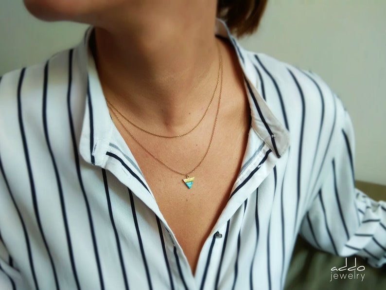 May include: A gold necklace with a small triangle pendant. The pendant is made of yellow, blue, and white enamel. The necklace is layered on a person wearing a white and black striped shirt.