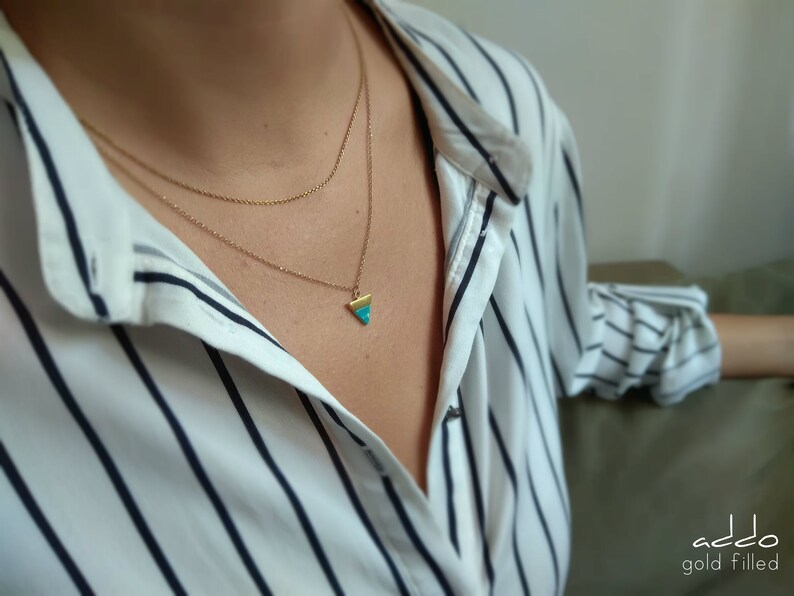 May include: A gold filled necklace with a turquoise triangle pendant. The necklace is layered on a model wearing a white and black striped shirt.
