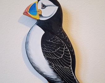 May include: A hand-painted wooden puffin with a black and white body, orange feet, and a colorful beak. The puffin is facing left and has a white background.