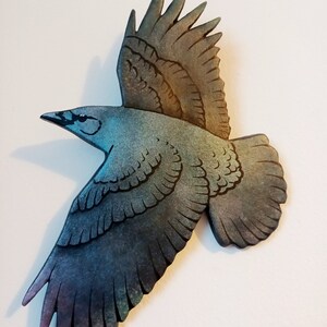 Può includere: Una scultura di uccello in legno dipinta in tonalità di blu, verde e viola. L'uccello è in volo con le ali spiegate. La scultura presenta un design dettagliato e una finitura testurizzata.