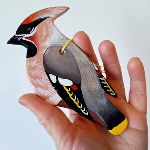 Könnte beinhalten: Handbemaltes Holzvogel-Ornament, das einen Seidenschwanz darstellt. Der Vogel ist in den Farben Rosa, Grau, Schwarz, Weiß, Gelb und Rot bemalt. Das Ornament hat eine Schnur zum Aufhängen.