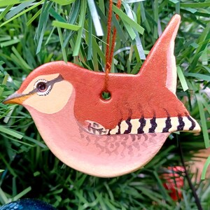 Könnte beinhalten: Ein handbemalter Keramik-Vogel-Anhänger mit braunen, rosa und schwarzen Details. Der Vogel sitzt auf einem Ast und hat ein Loch zum Aufhängen.