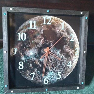 May include: Square wall clock with a black frame and a detailed moon phase design. The clock face features a realistic depiction of the moon's surface with white numerals and copper-colored hands. The frame is accented with small, turquoise-colored studs.