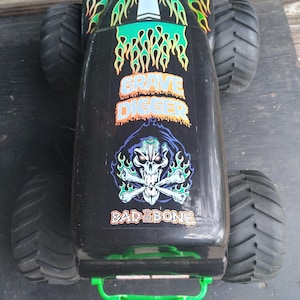Pu&ograve; includere: Monster truck giocattolo nero con pneumatici grandi. Il camion presenta le parole "Grave Digger" e "Bad to the Bone" con una grafica di teschio e ossa incrociate. Il camion ha disegni di fiamme verdi e gialle.