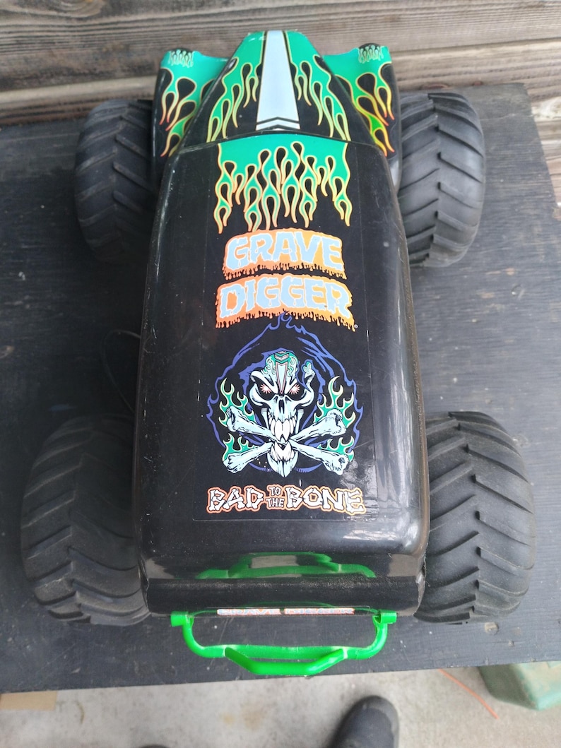 Pu&ograve; includere: Monster truck giocattolo nero con pneumatici grandi. Il camion presenta le parole "Grave Digger" e "Bad to the Bone" con una grafica di teschio e ossa incrociate. Il camion ha disegni di fiamme verdi e gialle.